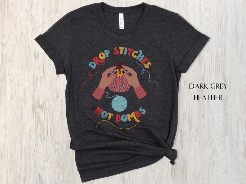 May include: Dark grey heather t-shirt with a colorful graphic. The graphic features hands knitting with the text "DROP STITCHES NOT BOMBS" in a circular design. A ball of blue yarn is also in the design.