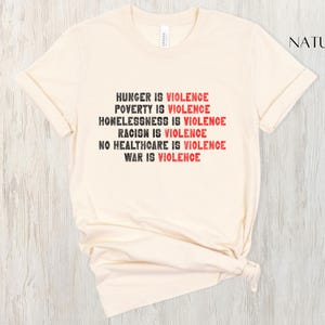 May include: A cream-coloured t-shirt with the words "HUNGER IS VIOLENCE, POVERTY IS VIOLENCE, HOMELESSNESS IS VIOLENCE, RACISM IS VIOLENCE, NO HEALTHCARE IS VIOLENCE, WAR IS VIOLENCE" printed in black and red. The shirt is made of soft material.