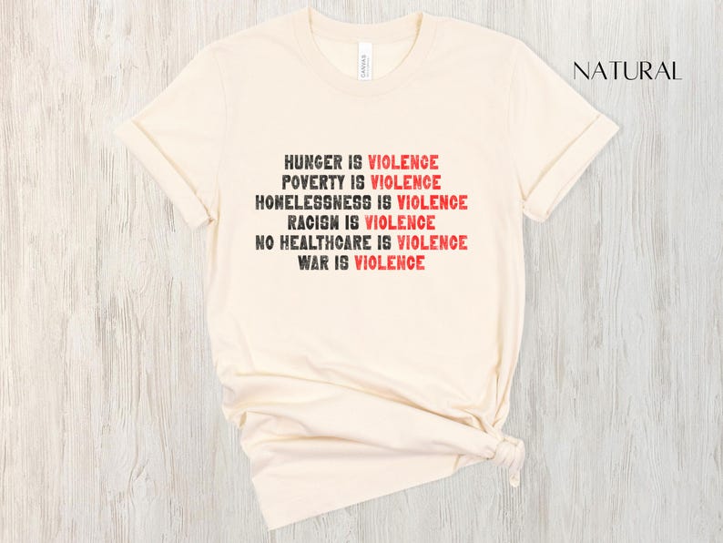 May include: A cream-coloured t-shirt with the words "HUNGER IS VIOLENCE, POVERTY IS VIOLENCE, HOMELESSNESS IS VIOLENCE, RACISM IS VIOLENCE, NO HEALTHCARE IS VIOLENCE, WAR IS VIOLENCE" printed in black and red. The shirt is made of soft material.