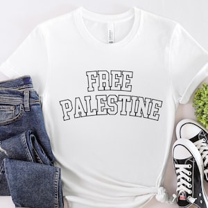 Może przedstawiać: Biała koszulka z czarnym napisem "FREE PALESTINE" w stylu czcionki uniwersyteckiej. Koszulka leży płasko na białym tle z parą czarnych butów sportowych i niebieskimi dżinsami.