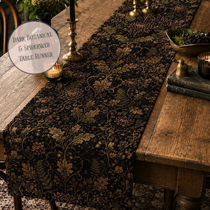 Könnte beinhalten: Ein langer, rechteckiger Tischläufer mit schwarzem Hintergrund und einem detaillierten botanischen Muster aus goldbraunen Eichenblättern, Farnen und feinen Spinnennetzmotiven. Der Text "DARK BOTANICAL & SPIDERWEB TABLE RUNNER" ist aufgedruckt.