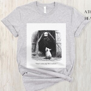 Puede incluir: Camiseta gris jaspeado con una ilustraci&oacute;n en blanco y negro de un cerdo mirando una puerta de granero. La puerta del granero tiene las palabras "FUCK ICE" escritas. El texto "That's some pig! He's radiant." est&aacute; debajo.