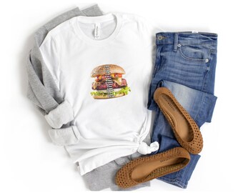 May include: A white t-shirt featuring a graphic of a cheeseburger with text layered over the patty, paired with a gray long-sleeve shirt, blue denim jeans, and tan woven flat shoes. The burger graphic includes lettuce, tomato, and cheese.