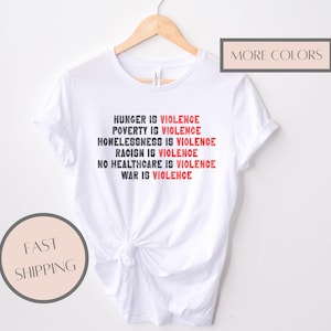 May include: White t-shirt with the text "HUNGER IS VIOLENCE, POVERTY IS VIOLENCE, HOMELESSNESS IS VIOLENCE, RACISM IS VIOLENCE, NO HEALTHCARE IS VIOLENCE, WAR IS VIOLENCE" in black and red. The shirt is displayed on a wooden hanger.