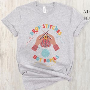 May include: Athletic heather t-shirt with a colorful graphic. The graphic features hands knitting with needles and yarn, with the words "DROP STITCHES NOT BOMBS" in a circular design. A ball of blue yarn is also included.