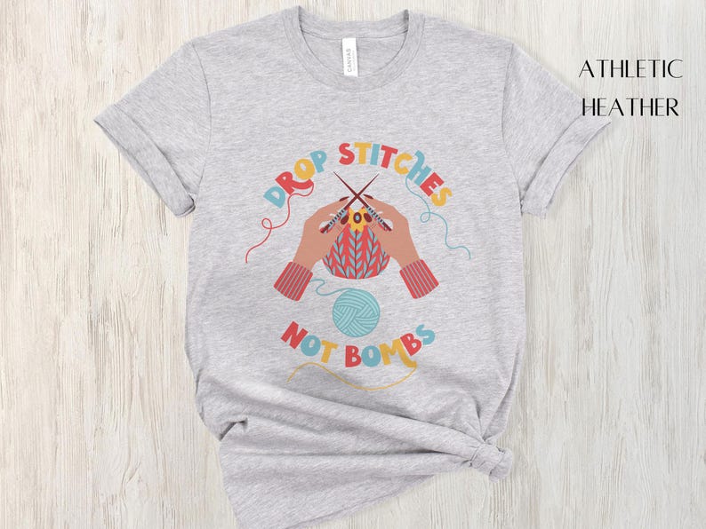 May include: Athletic heather t-shirt with a colorful graphic. The graphic features hands knitting with needles and yarn, with the words "DROP STITCHES NOT BOMBS" in a circular design. A ball of blue yarn is also included.