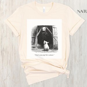 Puede incluir: Camiseta color crema con una ilustraci&oacute;n en blanco y negro de un cerdo mirando un granero. La puerta del granero tiene las palabras "FUCK ICE" encima del cerdo. El texto "That's some pig! He's radiant." est&aacute; debajo.