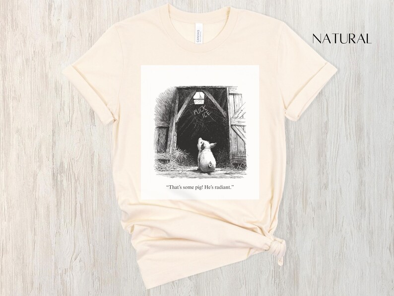 Puede incluir: Camiseta color crema con una ilustraci&oacute;n en blanco y negro de un cerdo mirando un granero. La puerta del granero tiene las palabras "FUCK ICE" encima del cerdo. El texto "That's some pig! He's radiant." est&aacute; debajo.