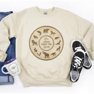Könnte beinhalten: Ein beiger Pullover mit einem braunen Kreis-Motiv, das verschiedene Tiere und den Text "All things serve the Beam" zeigt.