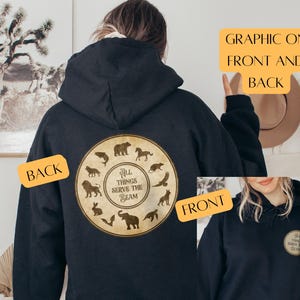 Könnte beinhalten: Schwarzer Kapuzenpullover mit einem kreisförmigen Aufdruck auf der Rückseite mit Tiersilhouetten und dem Text "ALL THINGS SERVE THE BEAM". Auf der Vorderseite befindet sich ein kleinerer, passender Aufdruck. Der Hoodie ist lässig.