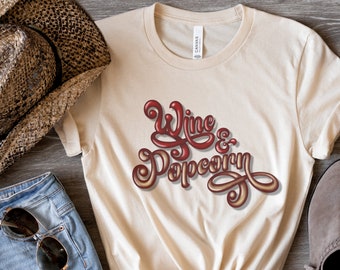 May include: A cream-colored short-sleeve t-shirt featuring the text Wine & Popcorn in a stylized, glossy red and gold script font. The shirt has a crew neckline and a standard fit, with the graphic centered on the chest area of the garment.