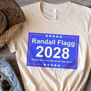 Puede incluir: Una camiseta beige con un gráfico rectangular azul y rojo que dice "Randall Flagg 2028" y "Shake the hand that shook the world."
