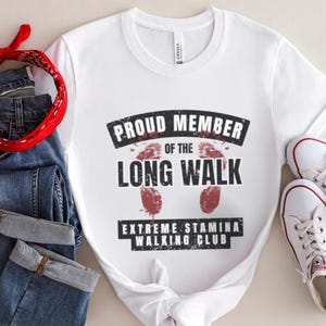 Op de afbeelding: Een wit t-shirt met de zwarte tekst PROUD MEMBER OF THE LONG WALK EXTREME STAMINA WALKING CLUB en twee rode voetafdrukken in het midden. Ook afgebeeld zijn een blauwe spijkerbroek, een rode bandana met een wit patroon en witte sneakers.