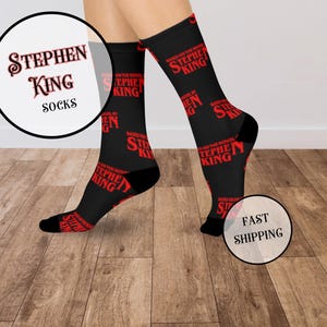 May include: Black socks with red text that reads "STEPHEN KING" and "BASED ON THE NOVEL OF STEPHEN KING." The socks are on a wooden floor. The text "FAST SHIPPING" is on a white circle.