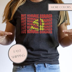 Może przedstawiać: Ciemnoszara koszulka z napisem "HOT COMMIE SUMMER" w czerwono-żółtym stylu retro. Obraz zawiera również napisy "MORE COLORS" i "FAST SHIPPING" w różowym polu.