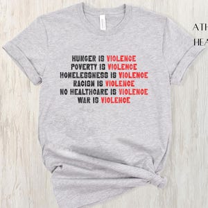 May include: Athletic heather grey t-shirt with the text "HUNGER IS VIOLENCE, POVERTY IS VIOLENCE, HOMELESSNESS IS VIOLENCE, RACISM IS VIOLENCE, NO HEALTHCARE IS VIOLENCE, WAR IS VIOLENCE" in black and red. The shirt is a crew neck.