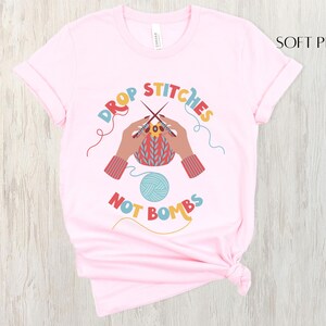 May include: Soft pink t-shirt with the text "DROP STITCHES NOT BOMBS" in colorful letters. The graphic shows hands knitting with needles and yarn. A ball of blue yarn is also featured in the design.