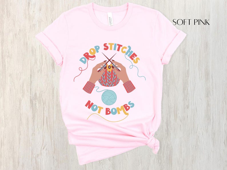 May include: Soft pink t-shirt with the text "DROP STITCHES NOT BOMBS" in colorful letters. The graphic shows hands knitting with needles and yarn. A ball of blue yarn is also featured in the design.