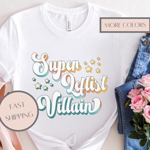 May include: White t-shirt with the text "Super Leftist Villain" in a retro font, accented with orange, teal, and yellow. The shirt is styled with blue jeans, pink heels, and a bouquet of pink roses.