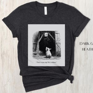 Puede incluir: Camiseta gris oscuro jaspeado con una ilustraci&oacute;n en blanco y negro de un cerdo mirando hacia un granero. La puerta del granero tiene las palabras "FUCK ICE". El texto "That's some pig! He's radiant." est&aacute; debajo.