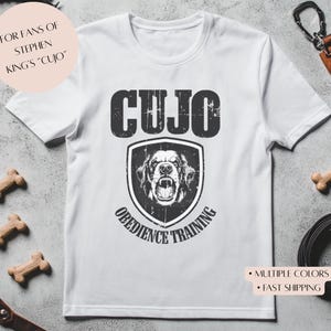 May include: White t-shirt with a black and white graphic of a dog's head in a shield with the text "CUJO OBEDIENCE TRAINING" and the text "FOR FANS OF STEPHEN KING'S "CUJO""