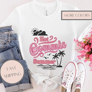 Może przedstawiać: Biała koszulka z różowo-czarną grafiką z napisem "Hot Commic Summer" z palmami, falami i ptakami. Koszulka jest połączona z niebieskimi dżinsami i białymi trampkami. Bukiet różowych kwiatów jest również na zdjęciu.