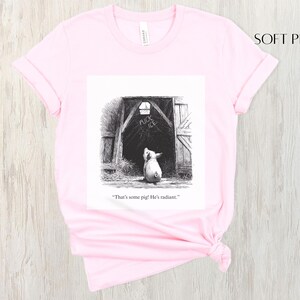 Puede incluir: Camiseta rosa claro con una ilustraci&oacute;n en blanco y negro de un cerdo mirando una puerta de granero. El texto de la ilustraci&oacute;n dice: "That's some pig! He's radiant."