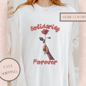 May include: A white t-shirt featuring a graphic of a hand holding a single red rose. Above the rose, the word "solidarity" is printed in a red, curved font, and below the rose, the word "Forever" is printed in the same red, stylised font.