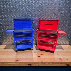 May include: Two rolling tool chests, one blue and one red, sit side-by-side on a wooden surface. Each chest has multiple drawers, a top compartment, and a side tray. The text "US GENERAL" is visible on the top of each chest.