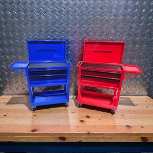 May include: Two small, rolling tool chests, one blue and one red, sit on a wooden surface. Each chest has multiple drawers and a top compartment. The text "US GENERAL" is visible on both chests.