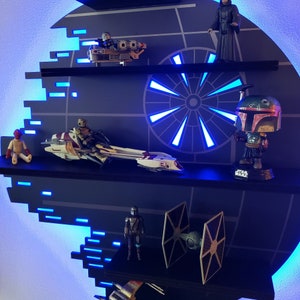 Op de afbeelding: Een Star Wars-thema rek met blauwe LED-verlichting. Het rek is gevormd als de Death Star en heeft verschillende Star Wars-personages en voertuigen, waaronder een speederbike, een TIE-fighter en een Boba Fett-figuur.