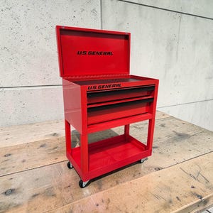May include: A red US GENERAL rolling tool chest with an open top and two drawers. The tool chest is on wheels and sits on a wooden surface. The text "US GENERAL" is printed on the top and front of the chest.