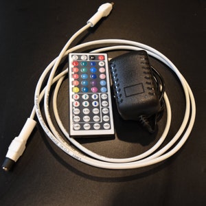 Op de afbeelding: Een wit snoer met een zwarte adapter en een afstandsbediening met verschillende gekleurde knoppen. De afstandsbediening heeft de tekst "DIY1, DIY2, DIY3, DIY4, DIY5, DIY6, JUMP, JUMP, FADE1, FADE1, SLOW, QUICK, LOW, AUTO, FLASH" erop.