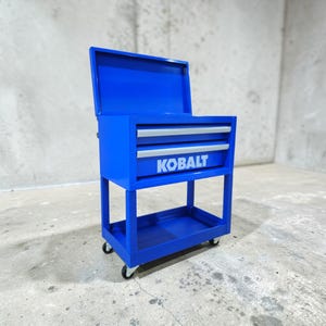 May include: A bright blue rolling tool chest with the lid open. The chest has two drawers with silver handles and the word "KOBALT" printed on the front. The tool chest is on wheels and has a bottom shelf.