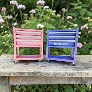 May include: Two rolling tool chests, one pink and one purple, each featuring three drawers with silver-toned handles and the white text KOBALT printed on the front. Both units are supported by four small caster wheels at the base.