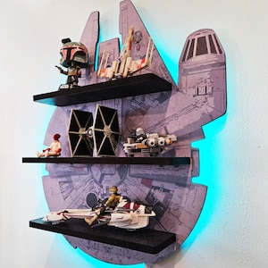 Peut inclure: Une étagère murale sur le thème de Star Wars avec trois étagères. L'étagère est en forme de vaisseau spatial Millennium Falcon. Les étagères sont remplies de jouets et de figurines Star Wars, dont un chasseur TIE, un X-Wing, une figurine Boba Fett et un speeder.