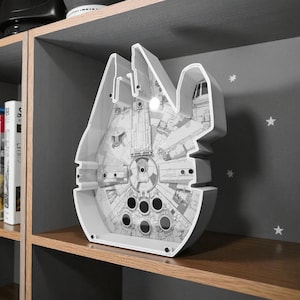 May include: A white, Star Wars-themed night light shaped like the Millennium Falcon. The light is displayed on a wooden shelf, with a detailed interior design visible. The light has a series of circular openings and a cockpit.