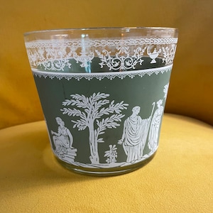 May include: A clear glass tumbler with a green base featuring white neoclassical figures and a tree design. The top rim has a white decorative pattern. The glass is cylindrical and sits on a yellow surface.