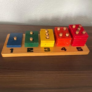 May include: A wooden educational toy with a light brown base and five colorful blocks. The blocks are blue, green, yellow, orange, and red, each with wooden pegs. Numbers 1 through 5 are printed below the blocks.