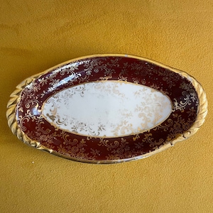May include: An oval-shaped ceramic dish with a burgundy and gold floral design. The dish has a white interior and a raised rim.