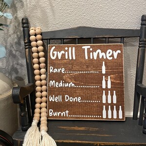 May include: A wooden sign with the text "Grill Timer" and a visual guide for different levels of doneness for grilled meat. The sign is displayed on a black rocking chair with a white tassel.