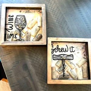 Op de afbeelding: Twee houten schaduwdozen met kurken stoppen erin. De eerste doos heeft een wijnglas en de tekst "wine time". De tweede doos heeft een kurkentrekker en de tekst "screw it".