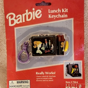 May include: A Barbie Lunch Kit keychain in original red packaging. The black lunch box features a graphic of a blonde figure and the Barbie logo. It includes a purple plastic clip, a brown thermos, and a yellow banana toy. Text includes Barbie, Lunch Kit Keychain, Ages 6 & Up, Really Works!, and Warning: Choking Hazard.