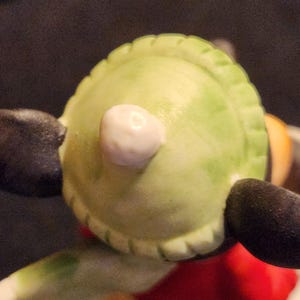May include: Close-up of a figurine's head, showing a light green hat with a white pom-pom. The figurine has black ears and a red and yellow outfit. The image is well-lit, highlighting the details of the ceramic figure.