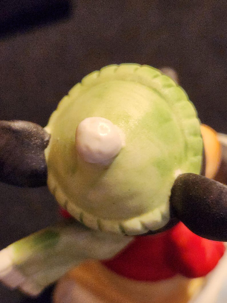 May include: Close-up of a figurine's head, showing a light green hat with a white pom-pom. The figurine has black ears and a red and yellow outfit. The image is well-lit, highlighting the details of the ceramic figure.
