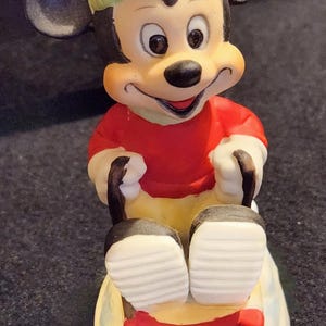 May include: A ceramic Mickey Mouse figure in a red top, green hat, and yellow trousers, sitting on a red and white sled. The figure has black ears and is smiling. A collectible item.