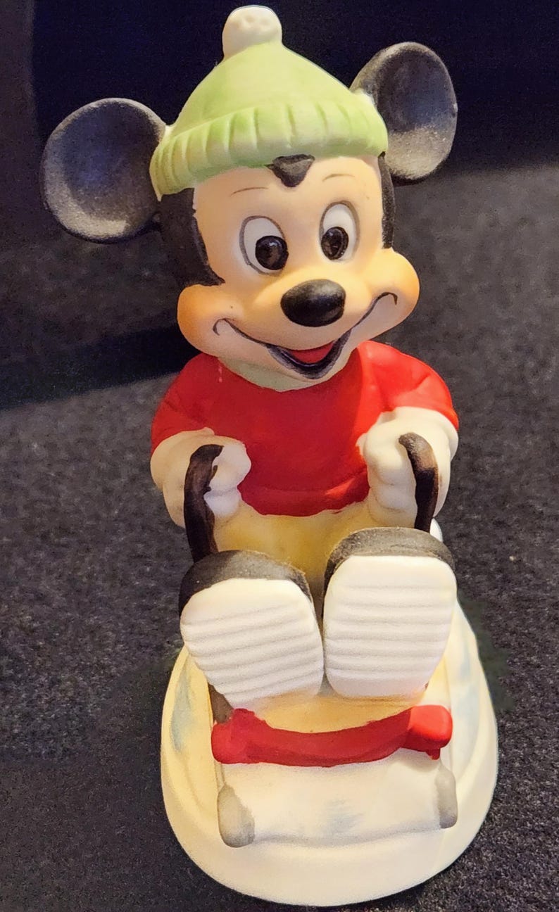 May include: A ceramic Mickey Mouse figure in a red top, green hat, and yellow trousers, sitting on a red and white sled. The figure has black ears and is smiling. A collectible item.