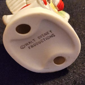 May include: A vintage ceramic figurine, featuring a white base with the text "WALT DISNEY PRODUCTIONS". The figurine has a red, yellow, and green design. The base has two holes.
