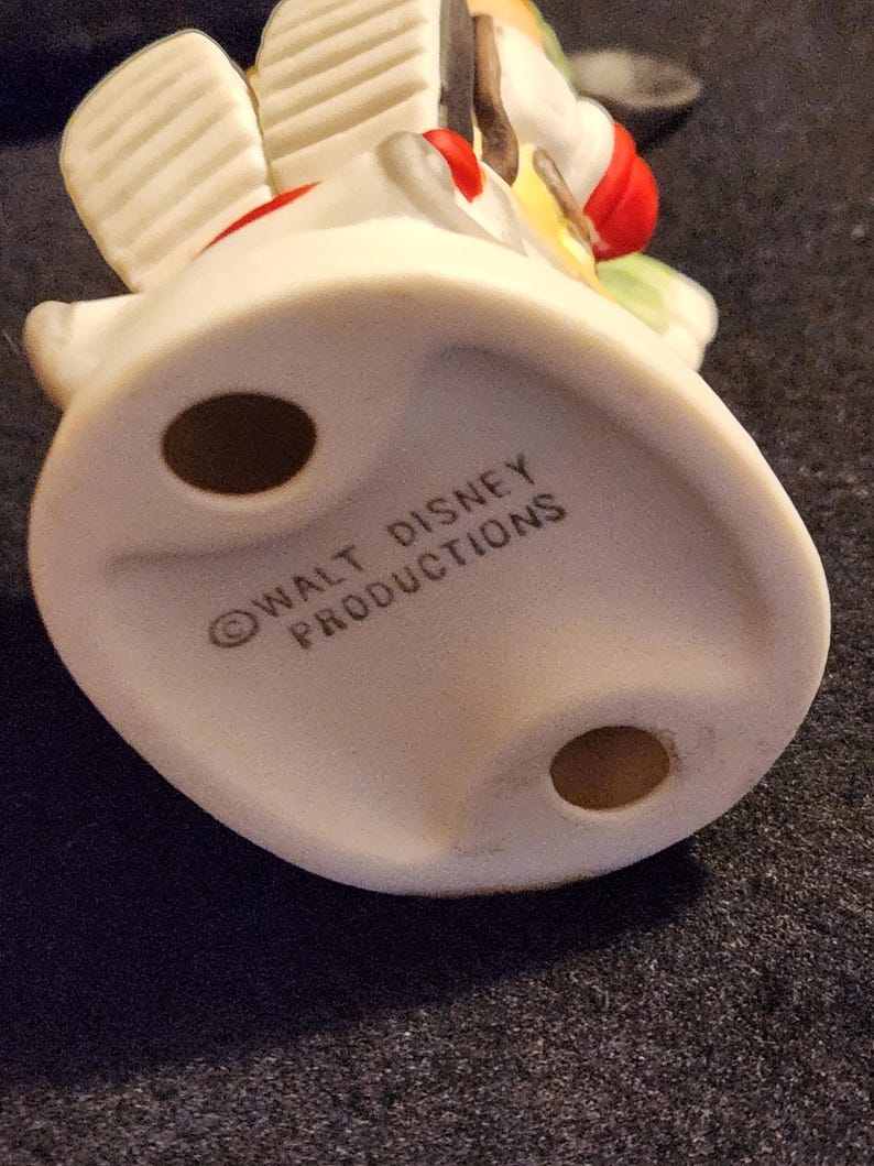 May include: A vintage ceramic figurine, featuring a white base with the text "WALT DISNEY PRODUCTIONS". The figurine has a red, yellow, and green design. The base has two holes.