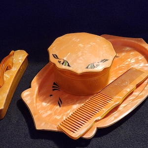 May include: A vintage peach-colored vanity set, including a rectangular brush, a round lidded container, a comb, and a decorative tray. The set has a marbled effect and black accents. The items are arranged on a dark surface.